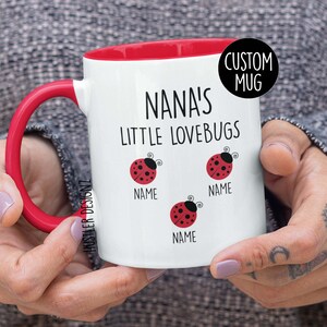 May include: White ceramic mug with a red interior and handle. The mug features the text "NANA'S LITTLE LOVEBUGS" and three ladybug illustrations. The word "NAME" is below each ladybug. A black circle with the words "CUSTOM MUG" is on the mug.