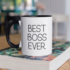 May include: White ceramic mug with a black handle and black rim. The mug has the text "BEST BOSS EVER." printed in black on the front. The mug is sitting on a stack of books with a floral pattern.