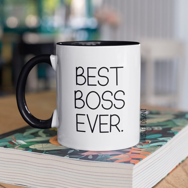 May include: White ceramic mug with a black handle and black rim. The mug has the text "BEST BOSS EVER." printed in black on the front. The mug is sitting on a stack of books with a floral pattern.