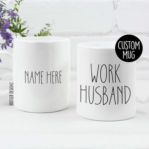 Op de afbeelding: Twee witte keramische mokken met zwarte tekst. De ene mok zegt "NAME HERE" en de andere mok zegt "WORK HUSBAND". De mokken staan op een wit oppervlak.