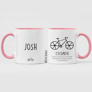 May include: Two white ceramic mugs with pink rims and handles. The mug on the left has the name "JOSH" printed on it, and a small bicycle illustration below. The mug on the right has a larger bicycle illustration and the word "CYCOPATH" printed below it. The text below the bicycle reads "A person suffering from chronic bike riding disorder with abnormal urges to ride and feel free."