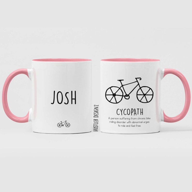 May include: Two white ceramic mugs with pink rims and handles. The mug on the left has the name "JOSH" printed on it, and a small bicycle illustration below. The mug on the right has a larger bicycle illustration and the word "CYCOPATH" printed below it. The text below the bicycle reads "A person suffering from chronic bike riding disorder with abnormal urges to ride and feel free."