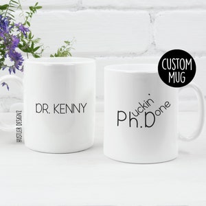 May include: Two white ceramic mugs. One mug says "Dr. Kenny" and the other mug says "Ph.D.  Fuckin' Done".
