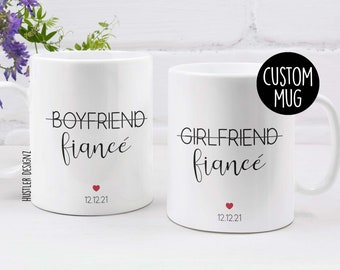Op de afbeelding: Twee witte keramische mokken met zwarte tekst. Op één staat "Boyfriend" doorgestreept en vervangen door "fiancé". Op de andere staat "Girlfriend" doorgestreept en vervangen door "fiancé". Elke mok heeft een rood hart en de datum 12.12.21.