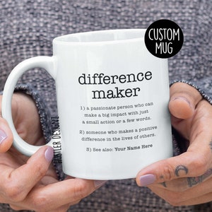 Pode incluir: Caneca de cerâmica branca com alça preta. A caneca tem um gráfico preto que diz "difference maker" e uma definição da palavra. A definição inclui o texto "Veja também: Seu nome aqui".