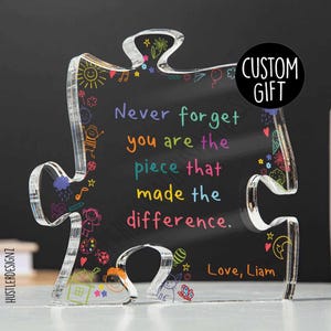 Op de afbeelding: Een transparant acryl puzzelstuk met een zwarte achtergrond en kleurrijke tekeningen. De tekst "Vergeet nooit dat je het stuk bent dat het verschil heeft gemaakt." staat in wit. De tekst "Love, Liam" staat rechtsonder.