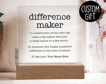 Puede incluir: Un letrero de acrílico transparente con la palabra "difference maker" impresa en negro. El letrero define a un creador de diferencia como una persona apasionada que puede tener un gran impacto con solo una pequeña acción o unas pocas palabras. El letrero también afirma que un creador de diferencia es alguien que hace una diferencia positiva en la vida de los demás. El letrero incluye el texto "Ver también: Tu nombre aquí". El letrero está sobre una base de madera y tiene una etiqueta "REGALO PERSONALIZADO" en la esquina superior derecha.