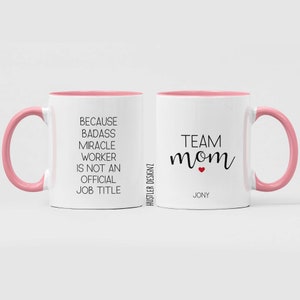 May include: Two white mugs with pink rims and handles. One mug says "Because badass miracle worker is not an official job title". The other mug says "Team mom" with a red heart and the name "Jony" underneath.