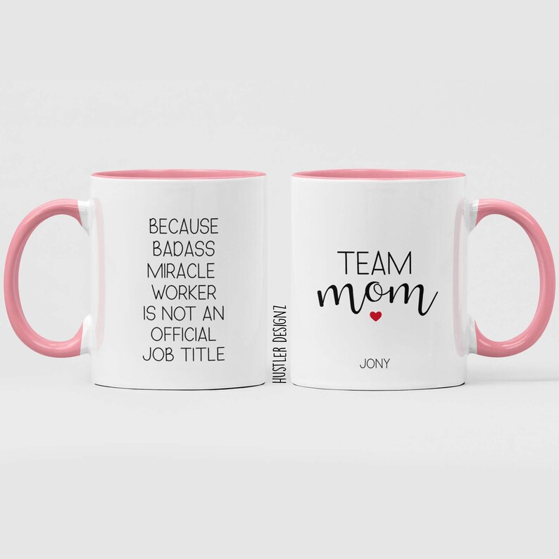 May include: Two white mugs with pink rims and handles. One mug says "Because badass miracle worker is not an official job title". The other mug says "Team mom" with a red heart and the name "Jony" underneath.