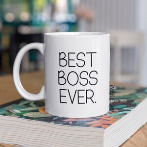 May include: White ceramic coffee mug with a black inscription that reads "BEST BOSS EVER." The mug is sitting on a stack of books with a floral design.