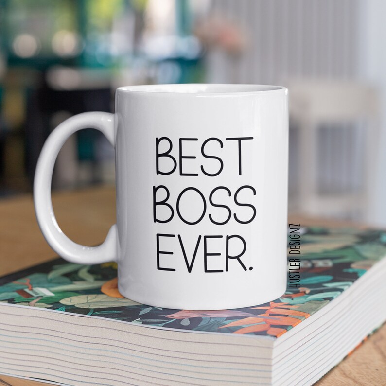 May include: White ceramic coffee mug with a black inscription that reads "BEST BOSS EVER." The mug is sitting on a stack of books with a floral design.