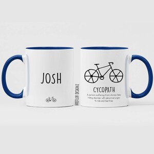 May include: White ceramic mug with a blue rim and handle. The mug has a black and white illustration of a bicycle and the text "CYCOPATH" with a definition below. The other side of the mug has the name "JOSH" in black text and a small bicycle illustration.