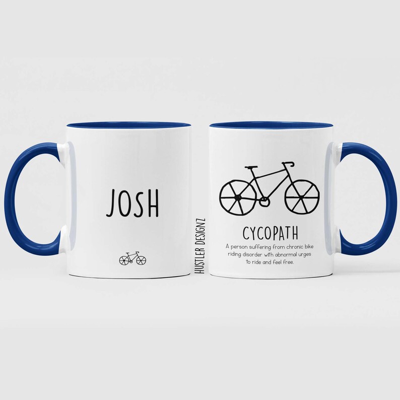May include: White ceramic mug with a blue rim and handle. The mug has a black and white illustration of a bicycle and the text "CYCOPATH" with a definition below. The other side of the mug has the name "JOSH" in black text and a small bicycle illustration.