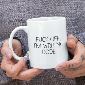 Puede incluir: Taza de cerámica blanca con el texto en negro "FUCK OFF. I'M WRITING CODE." La taza tiene un asa curva y la sostiene una persona.