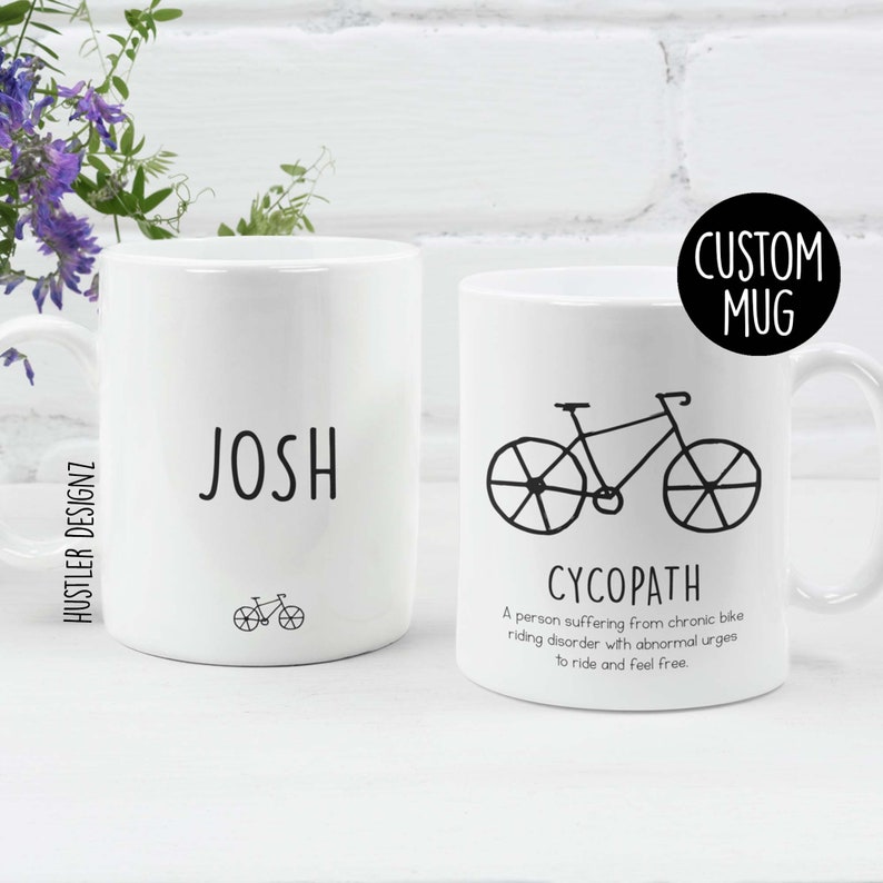 May include: Two white ceramic mugs with black text and a bicycle illustration. The mug on the left says "JOSH" and has a small bicycle illustration. The mug on the right says "CYCOPATH" and has a larger bicycle illustration. The text below the bicycle says "A person suffering from chronic bike riding disorder with abnormal urges to ride and feel free." The text "CUSTOM MUG" is in a circle at the top right of the image.