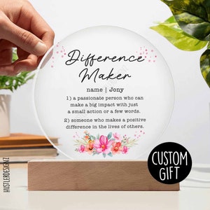 Puede incluir: Un letrero redondo de acrílico transparente con un diseño floral y el texto "Difference Maker" con el nombre "Jony" y una definición de la palabra. El letrero está sobre una base de madera y tiene una etiqueta de "Regalo personalizado".