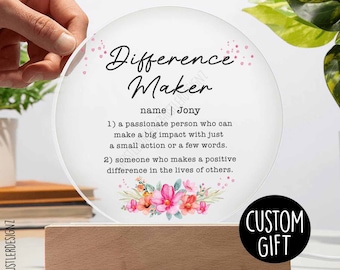 Pode incluir: Um sinal redondo de acrílico transparente com um design floral e o texto "Difference Maker" com o nome "Jony" e uma definição da palavra. O sinal está em uma base de madeira e tem um rótulo de "Presente personalizado".