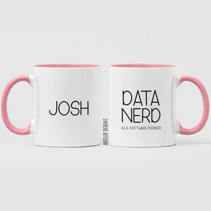 以下が含まれることがあります： ピンクの縁取りが付いた白いコーヒーカップが2つ。1つのカップには「Josh」と書いてあり、もう1つのカップには「Data Nerd AKA Software Engineer」と書いてあります。