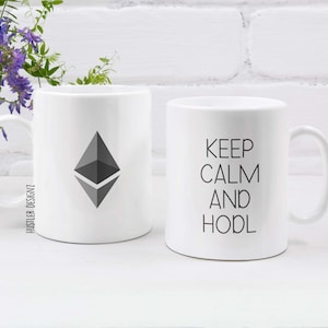 Könnte beinhalten: Zwei weiße Keramikbecher. Ein Becher zeigt ein graues Ethereum-Kryptowährungslogo. Der andere Becher hat den Text "Keep Calm and HODL" in Schwarz gedruckt.