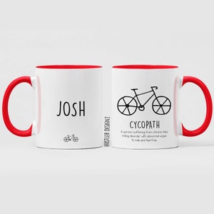 May include: White ceramic mug with a red rim and handle. The mug has a black illustration of a bicycle and the text "CYCOPATH" with a definition below. The mug also has the name "JOSH" on the other side.