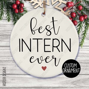 May include: A white ceramic ornament with the text "best intern ever" in black script. A small red heart is below the word "ever". The ornament has a gold string for hanging and a black circle with the words "custom ornament".