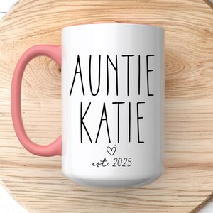 May include: White ceramic mug with a pink handle. The mug has black text that reads "Auntie Katie est. 2025" with a small heart symbol.