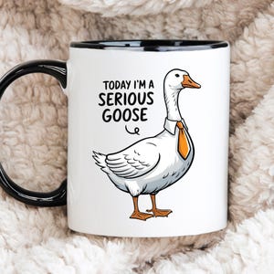Op de afbeelding: Witte keramische mok met een zwarte rand en handvat. De mok heeft een cartoon-gans met een stropdas en de tekst "Today I'm a Serious Goose".