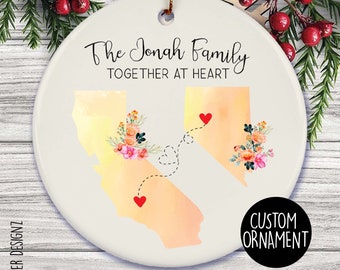 Puede incluir: Adorno navideño de cerámica blanca con el texto "The Jonah Family TOGETHER AT HEART". Presenta ilustraciones de acuarela de California y Nevada, conectadas por una línea de puntos con corazones. También hay detalles florales y las palabras "CUSTOM ORNAMENT".