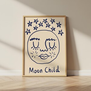 May include: A framed wall art print featuring a cream-colored background with a dark blue line drawing of a moon face with closed eyes and eyelashes. Above the moon are several hand-drawn stars, and the text Moon Child is written below.