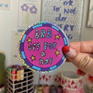Könnte beinhalten: Ein runder Aufkleber mit holografischem Rand und dem Text "BRB OFF FOR a NAP". Das Design umfasst einen rosa Hintergrund, blaue und gelbe Akzente. Der Aufkleber wird von einer Person gehalten.