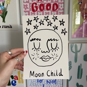 May include: A rectangular white art print featuring a black line drawing of a moon face with closed eyes, thick eyebrows, and a small mouth. Above the moon are several black stars, and the text "Moon Child" is written in black at the bottom.