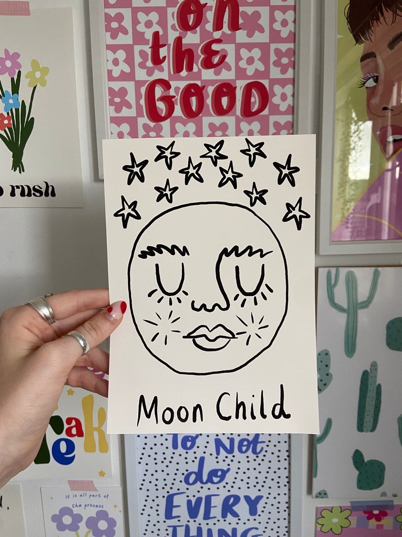 May include: A rectangular white art print featuring a black line drawing of a moon face with closed eyes, thick eyebrows, and a small mouth. Above the moon are several black stars, and the text "Moon Child" is written in black at the bottom.