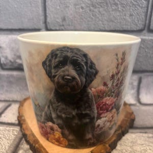 May include: A ceramic plant pot decorated with a portrait of a black dog with a curly coat. The pot has a floral design in pink and peach tones. It rests on a wooden, heart-shaped coaster.