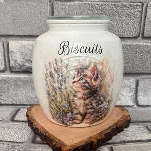 May include: A white ceramic biscuit jar with a textured surface, featuring a clear glass lid. The front displays a printed image of a brown tabby kitten sitting among purple lavender flowers, with the word Biscuits written in black script.