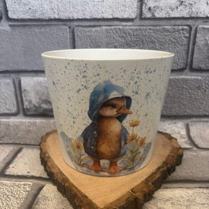 May include: A white ceramic plant pot featuring a duckling in a blue hooded jacket, surrounded by yellow flowers. The pot has blue paint splatters and rests on a wooden coaster. Ideal for small plants.