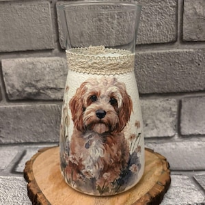 May include: A white ceramic mug with a black handle and the text "But First, Coffee" in a bold, black font. The mug is set against a blurred background. The mug appears to be approximately 10 cm tall.