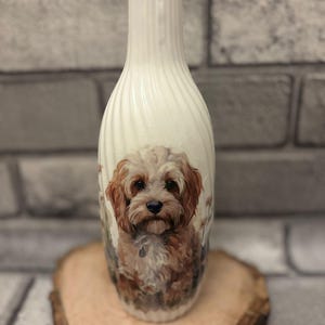 May include: A white ceramic vase featuring a painting of a brown and white dog. The vase has a textured, ridged design on the upper part. The vase is displayed on a wooden slice.