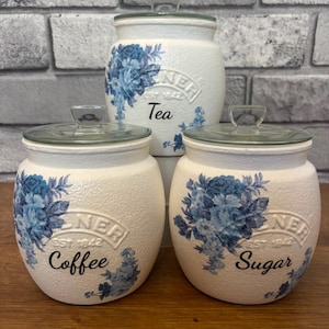 May include: Three off-white ceramic canisters with glass lids, featuring blue floral patterns. The canisters are labelled "Tea", "Coffee", and "Sugar". The text "EST 1842" is also present. The canisters have a textured finish and are designed for kitchen use.