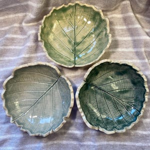 May include: Three ceramic leaf-shaped bowls in shades of green and blue-green. Each bowl has a detailed leaf vein pattern and a white rim. The bowls are arranged on a light purple, textured surface.
