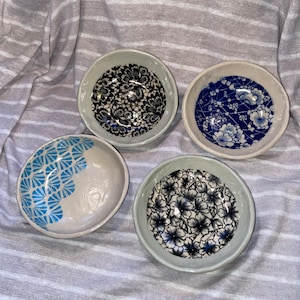 May include: Four small ceramic bowls with distinctive patterns. One bowl has a blue and white floral design, another a blue geometric pattern, and two others feature black and white floral designs. The bowls are displayed on a striped fabric background.