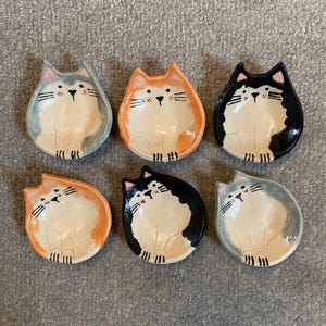 May include: Six ceramic cat-shaped dishes in various colours: blue, orange, and black. Each dish features a white cat design with black whiskers and a pink nose. The dishes are arranged on a grey surface.