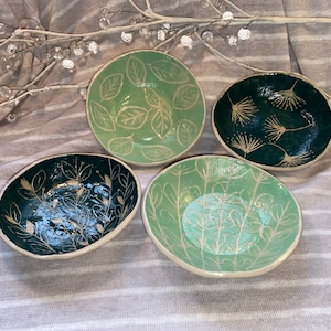 May include: Four small, handmade ceramic bowls in shades of green. Each bowl features a unique leaf or floral design in white. The bowls have a slightly irregular shape and a white rim, ideal for serving small portions or as decorative pieces.