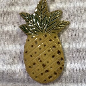 May include: A ceramic pineapple ornament with a textured surface and a glossy finish. The pineapple is mainly tan with dark brown spots and green leaves. The leaves have dark green detailing.