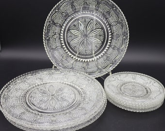 May include: Set of six clear glass dinner plates with a raised, embossed floral design. The plates are stacked in two piles, one with three plates and the other with three plates.