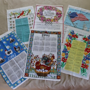May include: Six tea towels with calendar designs. The towels are white with colorful designs. The calendars are for the years 2000, 2001, 2002, 2004, and 2005. One towel has a red, white, and blue American flag design with the text "God Bless America".