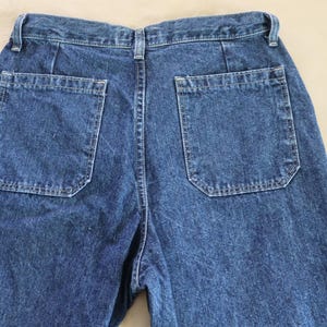 Op de afbeelding: Close-up van de achterkant van een blauwe spijkerbroek. De jeans heeft twee vierkante achterzakken met een unieke vorm. De denim heeft een medium wassing en de stiksels zijn zichtbaar. De jeans ligt plat op een neutrale ondergrond.