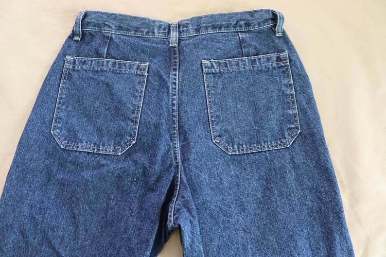 Op de afbeelding: Close-up van de achterkant van een blauwe spijkerbroek. De jeans heeft twee vierkante achterzakken met een unieke vorm. De denim heeft een medium wassing en de stiksels zijn zichtbaar. De jeans ligt plat op een neutrale ondergrond.