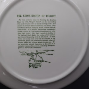 K&ouml;nnte beinhalten: Wei&szlig;e Keramikplatte mit gr&uuml;nem Text und einer Karte. Der Text lautet &bdquo;THE CROSS-ROUTES OF HISTORY&ldquo; und beschreibt die Geschichte von Hagerstown, Maryland. Eine Karte der Gegend ist ebenfalls enthalten.