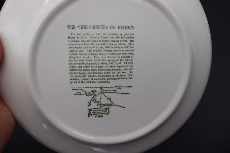 K&ouml;nnte beinhalten: Wei&szlig;e Keramikplatte mit gr&uuml;nem Text und einer Karte. Der Text lautet &bdquo;THE CROSS-ROUTES OF HISTORY&ldquo; und beschreibt die Geschichte von Hagerstown, Maryland. Eine Karte der Gegend ist ebenfalls enthalten.