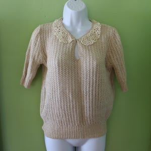 Puede incluir: Un suéter beige claro con un cuello de encaje decorativo. El suéter tiene mangas cortas y un escote de ojo de cerradura. La tela de punto tiene una apariencia texturizada. El suéter se exhibe en un maniquí blanco sobre un fondo verde.